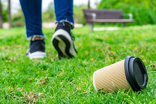 Low angle view of a disposable paper coffee cup with plastic lid discarded on the grass of a public park by walking away pedestrian. High resolution 42Mp studio digital capture taken with Sony A7rII and Sony FE 90mm f2.8 macro G OSS lens