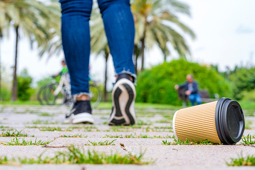 Low angle view of a disposable paper coffee cup with plastic lid discarded on the ground of a public park by walking away pedestrian. High resolution 42Mp studio digital capture taken with Sony A7rII and Sony FE 90mm f2.8 macro G OSS lens