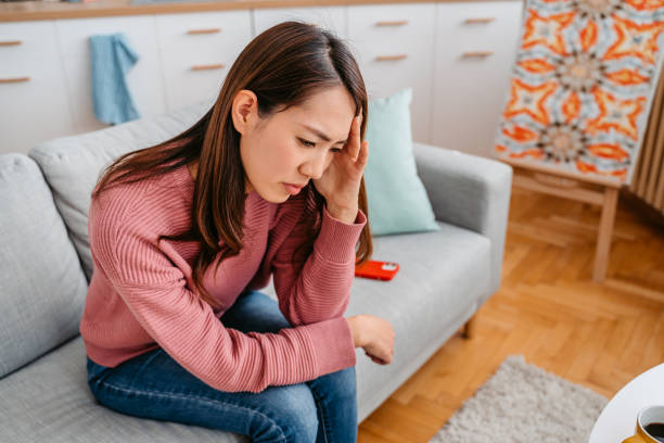 Young Woman Having A Headache Beautiful young Asian woman sitting on the sofa in the living room, having a headache. panic attack stock pictures, royalty-free photos & images