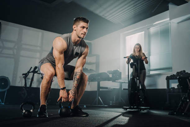 男性アスリートがケトルベルでトレーニ�ングをしている間、女性がヘルスクラブでエクササイズバイクでサイクリング - weightlifting crossfit weight training men ストックフォトと画像