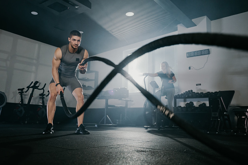 Young male athlete exercising with battle ropes while woman cycling on exercise bike at gym