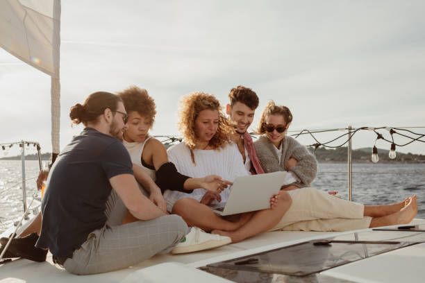 Friends enjoying a series on a streaming platform in the evening, sitting close to each other on boat deck of a sailboat, looking at a laptop stock photo