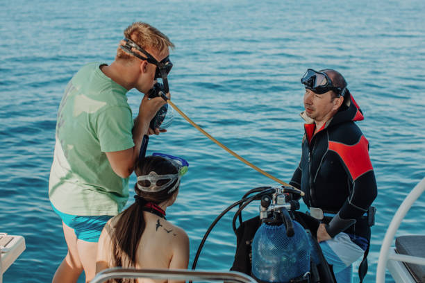 Young man with blond hair on a sailboat trying to breath through air regulator before diving in the sea, his dad and sister are watching him Young man with blond hair trying to breath through air regulator before diving in the sea, standing on boat deck of a sailboat, his dad and sister are watching him, calm sea in the background, steering wheel in the front scuba diving family stock pictures, royalty-free photos & images
