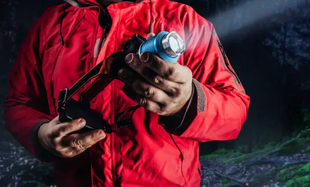 Photo of hiker man in red travel camping jacket holding shining head flashlight.