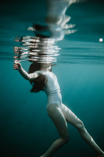 Woman with long brown hair and white swimsuit swimming in the sea, view from underwater, body underwater, head above water surface, calm sea