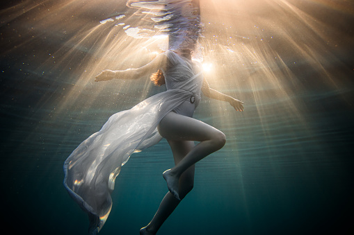 Young women with long brown hair and white dress swims in the sea, view from low angle underwater, sunlight coming from above, magical rays of light underwater