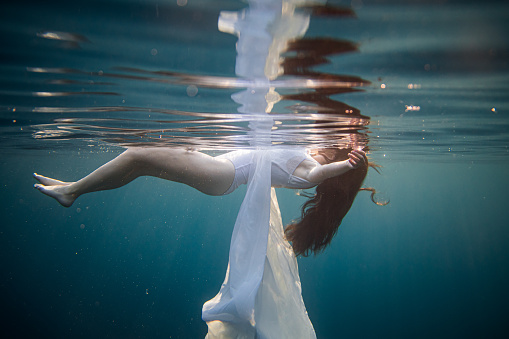 Women with long brown hair and white dress stretches her arms while floating on the water surface of the sea, view from the side from underwater