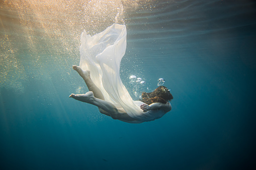 Women with long brown hair and white dress diving underwater in the sea, lots of huge air bubbles above her head after she jumped in the water, view from the side,