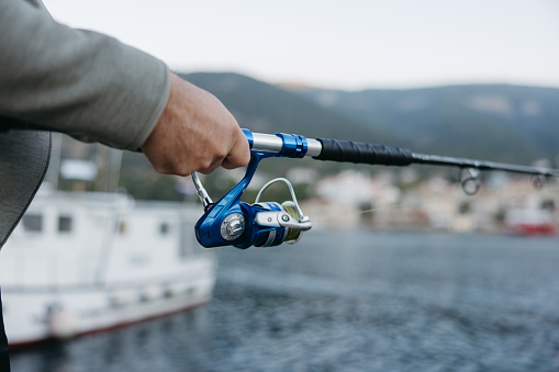 Close-up Fisherman holding a blue silver colored fishing rod with Fishing reel at the harbor, sea with island and village in the background, focus on hand and fisher rod, view from the side