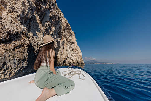 Woman with long brown hair and hat relaxing on front boat deck of a white ship on the sea, rope lying in front of her, coast with rock formations next to the ship, sea with island in the distance, rear view. Modra špilja, Biševo. Blue Cave Bisevo.