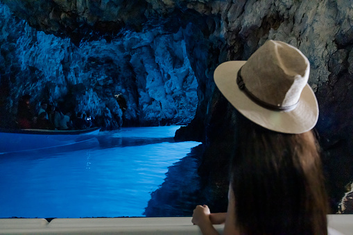 Woman with long brown hair and hat sitting on a boat, looking at a rock cave with perfectly clear blue water, rear view, upper body parts. Modra špilja, Biševo. Blue Cave Bisevo, Croatia.