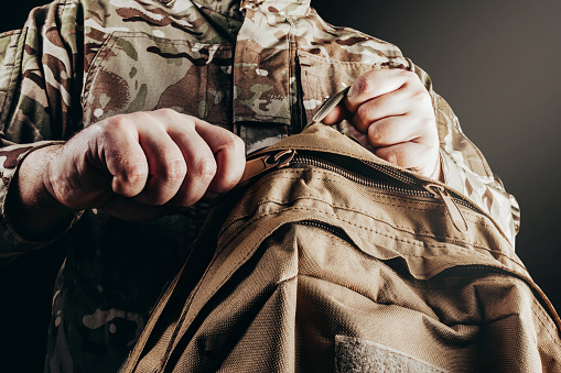 Photo of soldier in camouflaged uniform holding and unzipping backpack on black background.