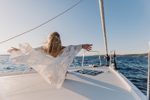 Woman with blond hair, wearing a swimsuit and white dress, stretching her arms out to feel the wind while sitting on front boat deck of a sailboat and looking at the sea, rear view