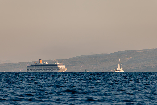 huge white colored cruise ship next to a large sailboat in front of an island with mountain range, view from distance, water with lots of small waves in front