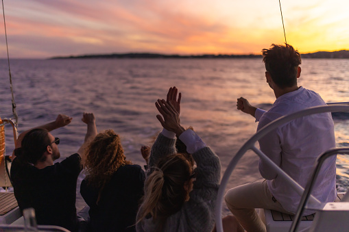 Group of friends sit on the back boat deck of a sailboat to watch the sunset, beautiful sunset sky above an island in the background, steering wheel in the front, cheerful mood, rear view