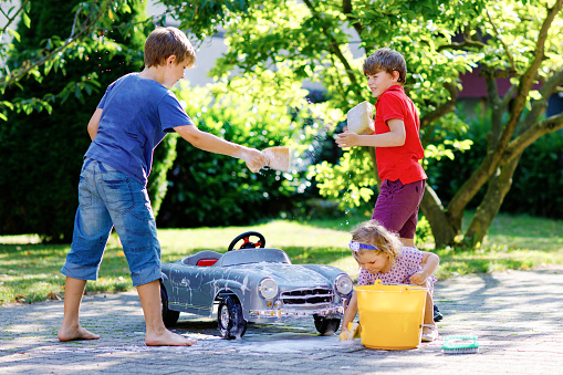 Three happy children washing big old toy car in summer garden, outdoors. Two boys and little toddler girl cleaning car with soap and water, having fun with splashing and playing with sponge