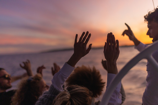 Friends watching the sunset together from boat deck of a sailboat while listening to music, raising arms in the sky, beautiful sunset sky above an island in the background, steering wheel in the front, cheerful mood