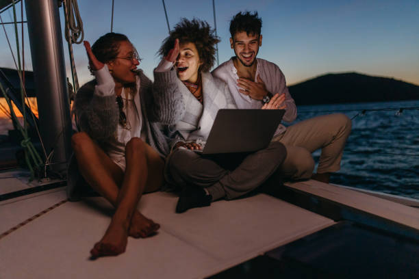 el grupo de amigos está emocionado después de reservar un viaje espontáneo para el día siguiente en una computadora portátil, sentado en la cubierta del barco de vela por la noche. - cheering business three people teamwork fotografías e imágenes de stock