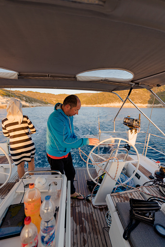 Adult couple standing on back boat deck of a sailboat underneath a roof, man with pullover and short brown hair controlling the steering wheel in the evening, women wearing a hand towel on her head and looking at the sea, drink bottles standing on a table in front, calm water with island in the background