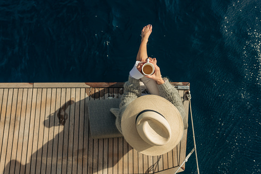 Women with beige hat holding a white cup of coffee in the morning, sitting on back wooden boat deck of a sailboat, bar feet legs are in the water, view from high angle