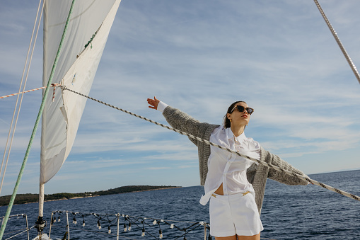 Woman with long brown hair and sunglasses stretches out arms while standing on boat deck of a sailboat to feel the wind, ropes of sail mast in the front, sea with island in the background