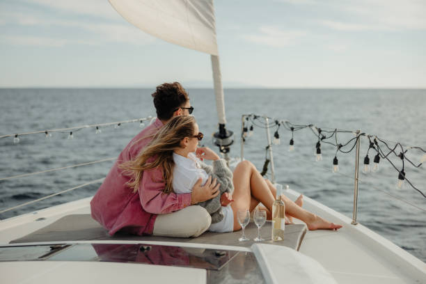 istock Man hugs his girlfriend while both sitting on front boat deck of a sailboat, wine bottle and glasses standing next to them 1392987513