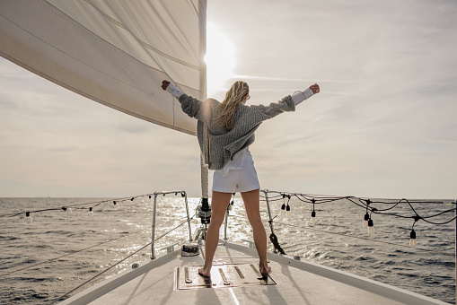 Woman with long blond brown hair and grey pullover spreads her arms while standing on front boat deck of a sailboat on the sea, feeling the wind, rear view, sun light behind her over the sea