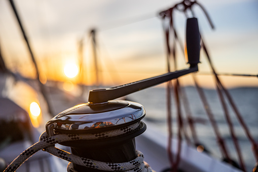 Close-up Winde of a sailboat with attached crank in the evening, rope holding the sail mast, beautiful sunset sky in the background, railing with ropes in the background, focus on forefront