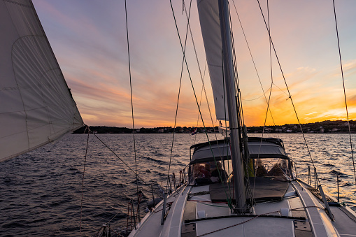 White colored large Sailboat on the sea with beautiful sunset sky in the background, view from front boat deck, people standing inside the cabin of the boat, calm sea with small waves and island in the background