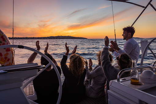 Group of friends, two men and three women, sitting on boat deck of a sailboat in the evening looking at a beautiful sunset above an island on the sea, clapping their hands to music, cheerful mood