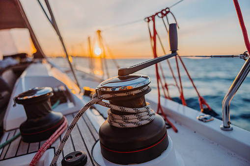 Winde of a sailboat with attached crank in the evening, rope holding the sail mast, empty boat deck, sea with island and beautiful sunset sky in the background