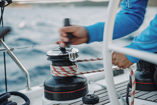Sailor man with blue pullover turns a crank Winde to retract the sail, sitting on boat deck of a sailboat on the sea in the evening, blurred motion, holding the rope with his hand