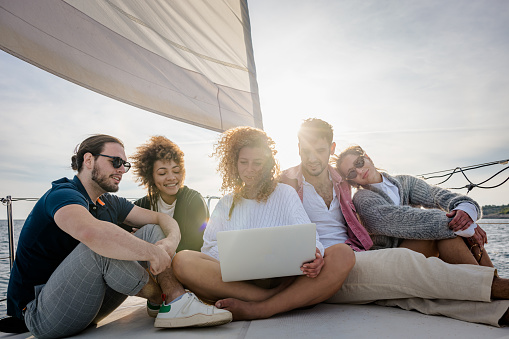 Friends, two men and three women, looking for travel destinations on their laptop while sitting close to each other on front boat deck of a sailboat in the evening, sunlight above their heads