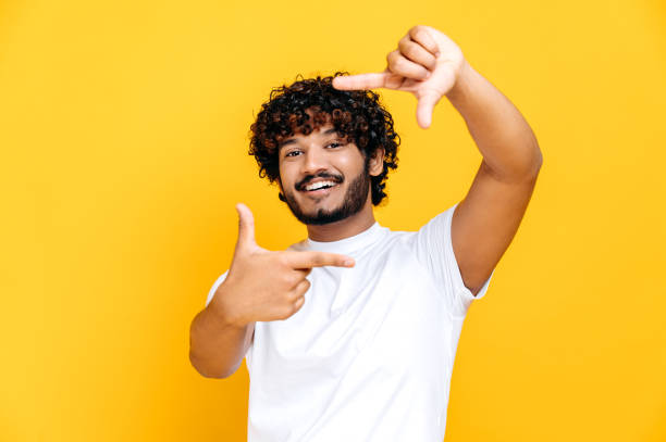 joyeux joyeux positif indien ou arabe millénaire gars en t-shirt blanc de base, faisant cadre imaginaire de caméra prenant des photos, debout sur un fond orange isolé, regarde la caméra, souriant - hommes photos photos et images de collection