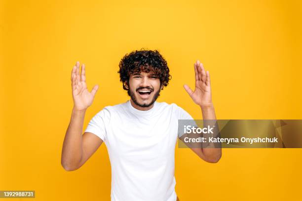 Confused Puzzled Indian Or Arabian Attractive Curlyhaired Young Man Emotionally Gesturing With His Hands Looking Inquiringly At The Camera Standing On An Isolated Orange Background Stock Photo - Download Image Now