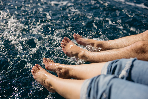 Man and woman putting their barefoot legs in the water while sitting next to each other on boat deck of a sailboat on the sea, women in the front wearing jeans shorts, close-up, view from the side, only legs