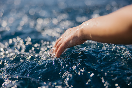 Woman's hand touching the blue water surface of the sea, lots of small waves around her hand, close-up of arm with white skin and hand touching the water