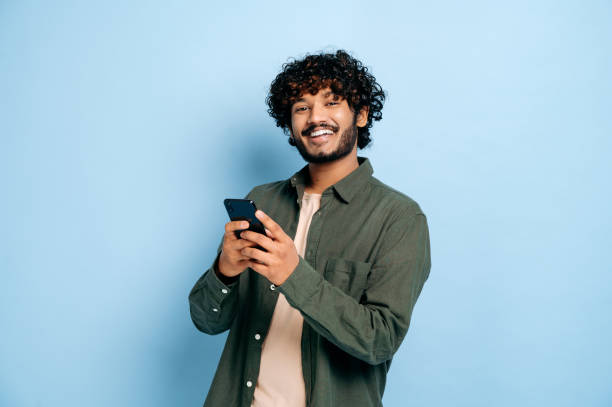 istock Happy handsome amazed stylish indian or arabian guy, using smartphone, chatting online, texting message, browsing internet, social media, looks at camera, stands on isolated blue background, smiling 1392990094