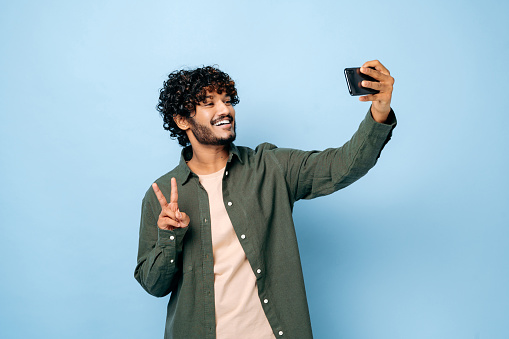 Cheerful happy indian or arabian excited guy with gladden face expression, in casual stylish clothes, doing selfie shot on smartphone and smiles into the camera, stands on isolated blue background