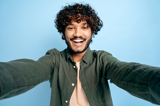 Cheerful Indian funny curly-haired guy with gladden face expression, in casual shirt, doing selfie shot on smartphone and smiling into the camera, stands on isolated blue background