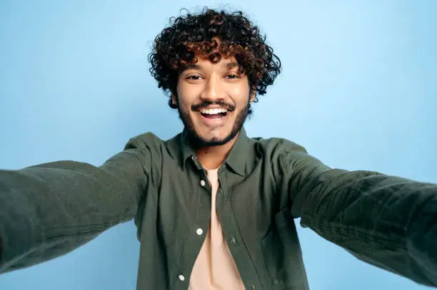 Photo of Cheerful Indian funny curly-haired guy with gladden face expression, in casual shirt, doing selfie shot on smartphone and smiling into the camera, stands on isolated blue background