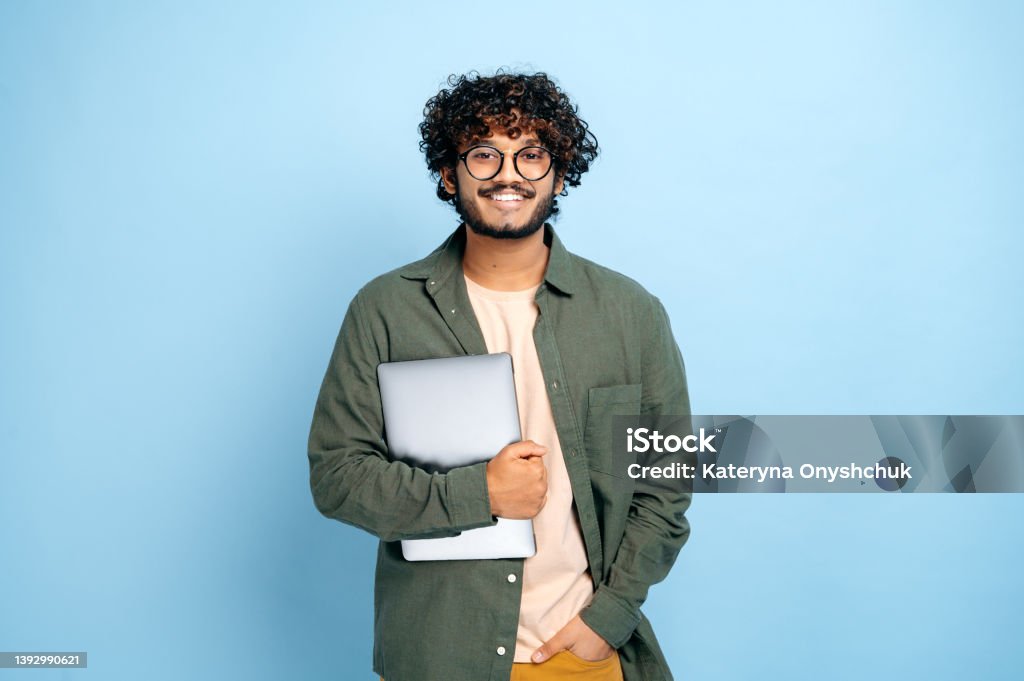 Smart handsome positive indian or arabian guy, with glasses, in casual wear, student or freelancer, holding a laptop in hand, standing on isolated blue background, looking at camera, smiling friendly Student Stock Photo
