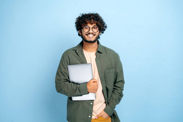 Smart handsome positive indian or arabian guy, with glasses, in casual wear, student or freelancer, holding a laptop in hand, standing on isolated blue background, looking at camera, smiling friendly stock photo