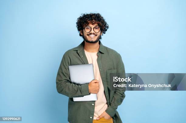 Smart Handsome Positive Indian Or Arabian Guy With Glasses In Casual Wear Student Or Freelancer Holding A Laptop In Hand Standing On Isolated Blue Background Looking At Camera Smiling Friendly Stock Photo - Download Image Now