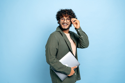 Smart handsome positive indian or arabian guy, with glasses, student, freelancer, creative designer, holding a laptop in hand, standing on isolated blue background, looking at camera, smiling friendly