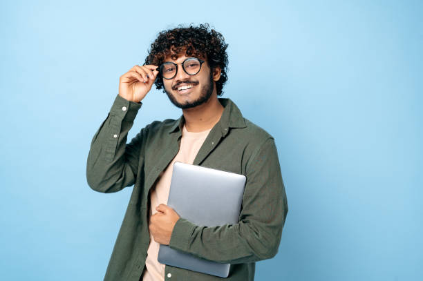 Smart handsome positive indian or arabian millennial guy, with glasses, student or freelancer, holding a laptop in hand, standing on isolated blue background, looking at the camera, smiling friendly stock photo