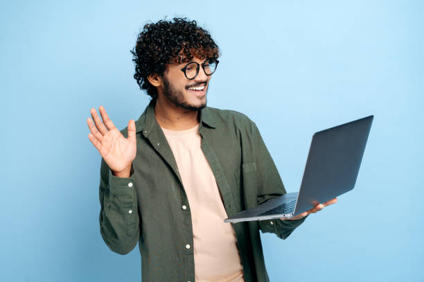 istock Online video communication. Indian or Arabic guy, in casual clothes, freelancer or student, uses a laptop for a video conference, stands on isolated blue background, greets an interlocutor, smiling 1392990632