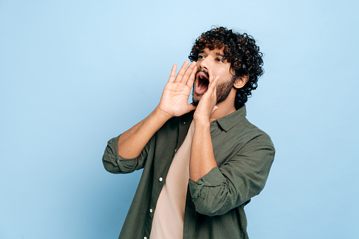 Excited indian or arabic curly haired guy in casual clothes, shouting loudly holding his hands near his mouth, making an announcement, calling out, standing on an isolated blue background