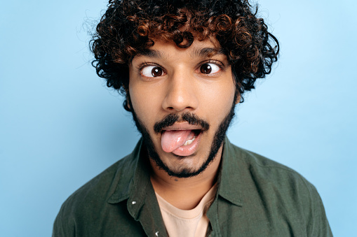 Close up face of indian or arabian curly funny guy, making a funny face, tongue out, squinted his eyes, standing on isolated blue background. Grimaces, fooling around
