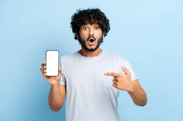 istock Shocked amazed indian or arabian guy, hold in hand cellphone with white empty mock-up screen for presentation, points finger at it, surprised looks at camera, stand on isolated blue background, smile 1392990643